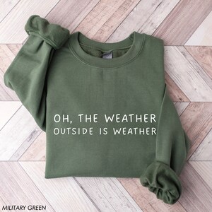 May include: Military green sweatshirt with the text "OH, THE WEATHER OUTSIDE IS WEATHER" in white. The sweatshirt is folded and has a crew neck and long sleeves. The words "MILITARY GREEN" are at the bottom.