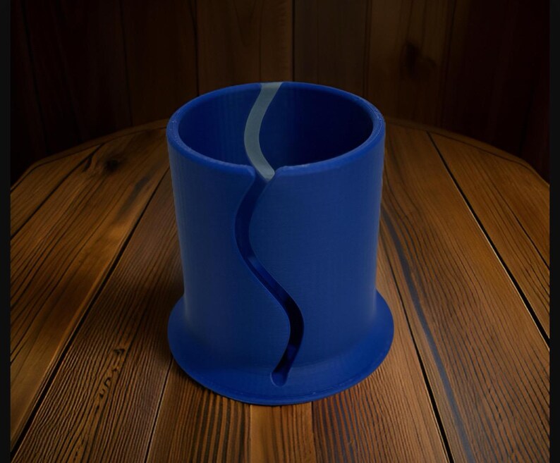Op de afbeelding: Een cilindrische blauwe houder met een doorlopende, verticale, S-vormige uitsparing aan de zijkant. Het object heeft een uitlopende voet en een gladde, matte afwerking, met een lichtgrijze binnenvoering zichtbaar door de opening in de voorwand.