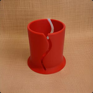 Op de afbeelding: Een cilindervormig rood object met een brede, uitlopende basis en een verticale, golvende gleuf die vanaf de bovenrand naar beneden loopt. De binnenkant van de cilinder is hol en het oppervlak is glad en mat met een witte accentkleur.