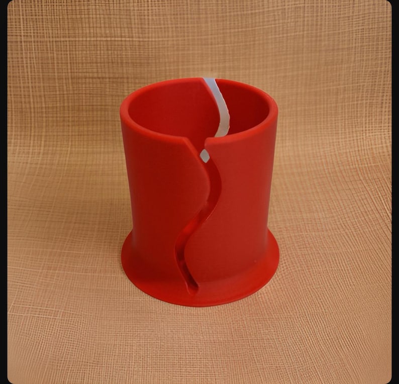 Op de afbeelding: Een cilindervormig rood object met een brede, uitlopende basis en een verticale, golvende gleuf die vanaf de bovenrand naar beneden loopt. De binnenkant van de cilinder is hol en het oppervlak is glad en mat met een witte accentkleur.