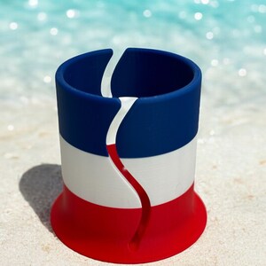May include: A red, white, and blue silicone cup holder with a split design. The cup holder is sitting on a white sandy beach.