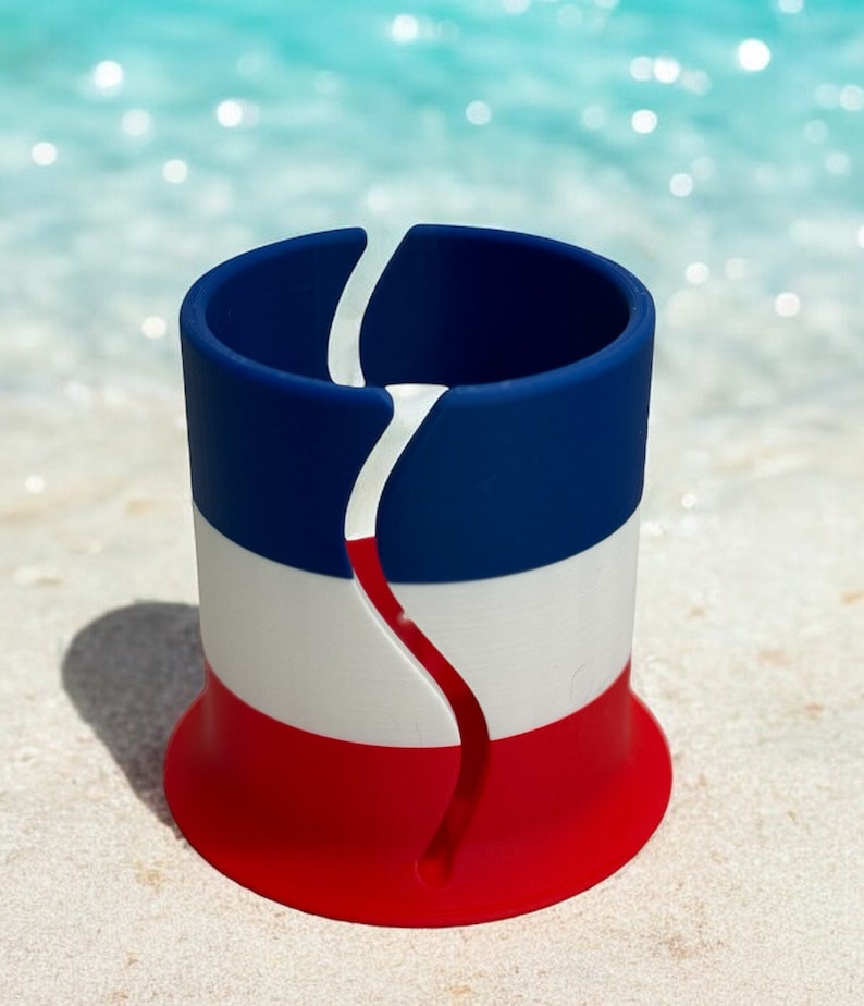 May include: A red, white, and blue silicone cup holder with a split design. The cup holder is sitting on a white sandy beach.