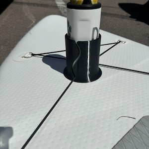 Op de afbeelding: Een zwarte cilindrische bekerhouder vastgezet met zwarte elastische koorden op een wit getextureerd oppervlak van een paddleboard. De houder bevat een wit drankblikje met een geel en zwart grafisch ontwerp en een pootafdruk-icoon.