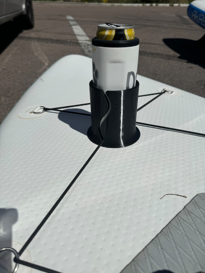 Op de afbeelding: Een zwarte cilindrische bekerhouder vastgezet met zwarte elastische koorden op een wit getextureerd oppervlak van een paddleboard. De houder bevat een wit drankblikje met een geel en zwart grafisch ontwerp en een pootafdruk-icoon.