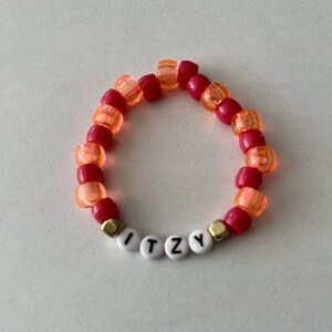May include: A beaded bracelet with red, orange, and gold beads. The bracelet spells out the word "ITZY" with white letter beads. The bracelet is on a white surface.
