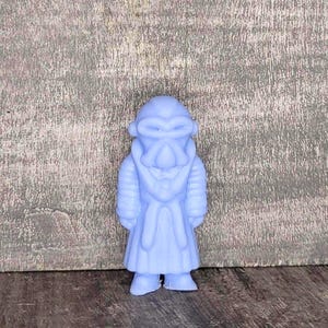 May include: A light blue figurine of a character with a mask-like face, wearing a long coat. The figurine has a detailed design and stands upright. The background is a textured, gray surface.