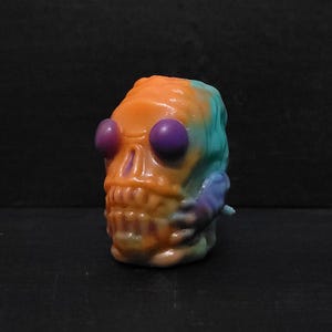 May include: A colorful skull-shaped figurine with large purple eyes. The skull is painted with orange, blue, and purple hues. The figurine has a detailed skeletal structure and is set against a dark background.