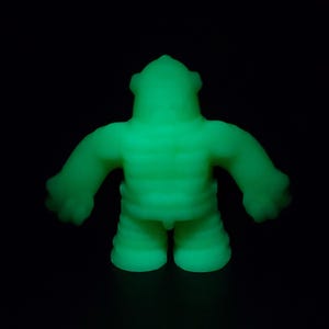 May include: A glowing green vinyl figure of a stylized humanoid creature. The figure has a textured, ribbed torso, short legs, and outstretched arms with rounded hands. The head has a distinctive, ridged crown. The figure is set against a black background.