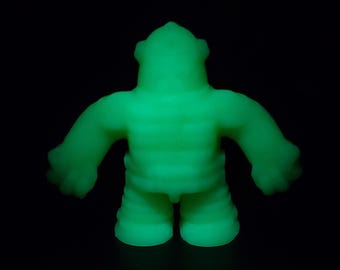 Può includere: Una figura in vinile verde brillante di una creatura umanoide stilizzata. La figura ha un torso strutturato e a coste, gambe corte e braccia tese con mani arrotondate. La testa ha una caratteristica corona a coste.