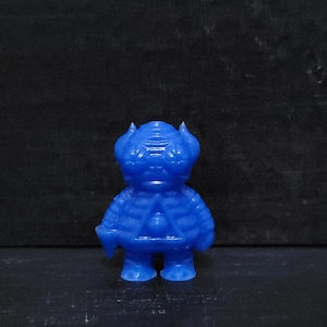 May include: A blue figurine of a stylized humanoid figure with a textured surface. The figure has two small horns and a triangular design on its chest. The figurine is standing on a dark surface.