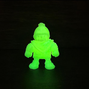 May include: A glowing green figurine of a stylized character with a hat, scarf, and chunky boots. The figure is brightly illuminated against a dark background, giving it a vibrant, almost neon appearance. The toy has a unique, cartoonish design.