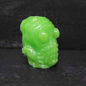 May include: A glowing green skull-shaped figurine with a textured surface. The figurine has prominent eye sockets and a detailed skeletal structure. The overall design suggests a spooky or Halloween theme.