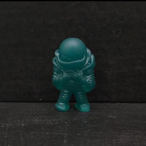 May include: A teal-colored astronaut figurine. The figurine is standing upright with its hands clasped in front. The astronaut is wearing a spacesuit with a helmet. The background is a dark, solid color.