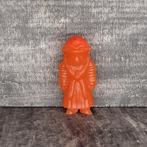 May include: An orange figurine of a person wearing a long coat and hat. The figurine has a detailed design and stands upright. The figure is made of a translucent material.