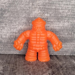 May include: Orange, stylized action figure with a textured surface. The figure has a helmet-like head, a rectangular torso, and thick arms and legs. The figure's design is reminiscent of a vintage toy or collectible.