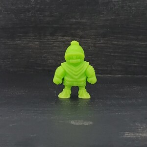 May include: A small, lime green figurine of a humanoid character with a helmet and armor-like details. The figure has a textured surface and stands on a dark background. The design is reminiscent of a vintage toy or collectible.