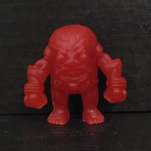 May include: A small, solid red monster figurine with a textured surface. The figure has a large head, muscular arms, and stubby legs. The face has a menacing expression, with a wide mouth and furrowed brow. The toy is made of a solid material.