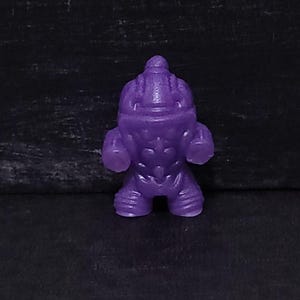 May include: A small, purple figurine of a muscular humanoid figure. The figurine has a detailed design with defined muscles, a helmet-like head, and large, rounded fists. The figure is standing upright.