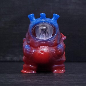 May include: A small, colorful figurine of a creature with a large, clear, bubble-like head. The figurine is primarily red with blue accents on the head and arms. The creature has three blue protrusions on top of its head.