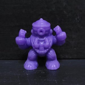 May include: A small, solid purple action figure of a humanoid robot with a bulbous head, large feet, and raised fists. The figure has a detailed chest and a small, circular opening on its head. The toy is standing on a dark surface.