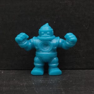 May include: A small, turquoise-colored figurine of a robot-like character with its fists clenched. The figure has a round head with a pointed top, a circular design on its chest, and is standing with its legs apart. The toy is made of a solid, smooth material.