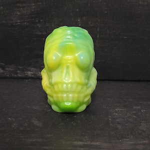 May include: A green and yellow skull-shaped figurine. The skull has large eye sockets, a prominent jaw, and a textured surface. The colors blend from green at the top to yellow at the bottom. The figurine is a unique decorative item.