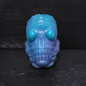 May include: A blue and purple skull-shaped figurine with large, round, teal-colored eyes. The skull has a detailed, textured surface and a wide, toothy grin. The color gradient shifts from blue to purple.
