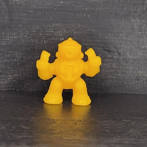 May include: A solid yellow action figure of a humanoid robot with a single eye and raised fists. The toy has a rounded head, thick arms and legs, and a detailed torso. The figure is standing on a dark surface.