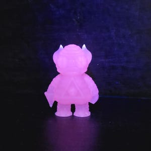 May include: A pink, translucent figurine of a stylized creature with horns and a triangular design on its chest. The figure has a rounded body, small legs, and is illuminated with a purple light against a dark background.