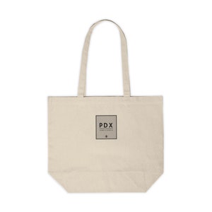 May include: A beige canvas tote bag with black handles and a black square logo that says "PDX Portland International Airport 45.5889° N 122.5937° W" with a small airplane symbol below the text.