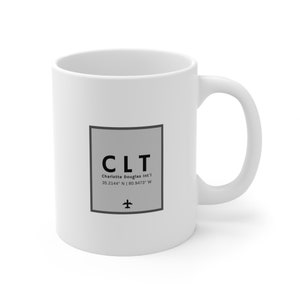 Könnte beinhalten: Weiße Keramiktasse mit einem schwarzen und grauen Design, das den Flughafencode "CLT" für den Charlotte Douglas International Airport sowie den Namen des Flughafens und die Koordinaten 35.2144° N | 80.9473° W zeigt.