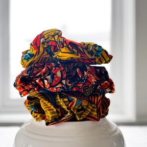 Peut inclure: Une pile de chouchous en tissu aux motifs colorés, avec des motifs rouges, jaunes et bleus. Les chouchous sont empilés sur un bol en céramique blanc. L'arrière-plan est une fenêtre lumineuse.