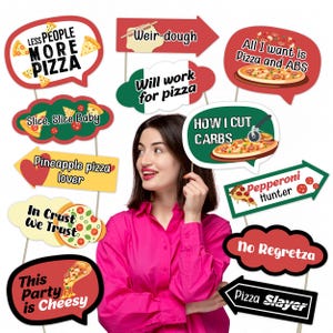May include: A woman in a pink shirt surrounded by pizza-themed photo booth props. The props include speech bubbles and arrows with phrases like "Less People More Pizza," "Slice, Slice Baby," and "Pizza Slayer." The props are in various colors and shapes.