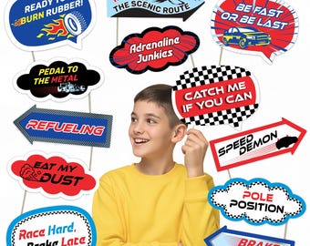 May include: A collection of racing-themed photo booth props on sticks. The props feature phrases like "Ready to Burn Rubber!", "Catch Me If You Can", and "Pole Position". The props are in various shapes, including speech bubbles and arrows, with bold colors and racing-related graphics.