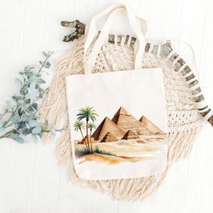 May include: Cream-colored tote bag featuring a watercolor illustration of the pyramids and palm trees. The bag has long, off-white handles and is made of a sturdy fabric. The design evokes an Egyptian theme.