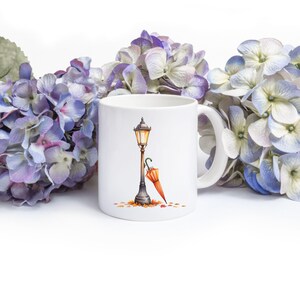 Pode incluir: Caneca de cer&acirc;mica branca com uma ilustra&ccedil;&atilde;o em aquarela de um lampi&atilde;o vintage, um guarda-chuva laranja e folhas de outono. A caneca &eacute; cercada por flores de hort&ecirc;nsia roxas e azuis.