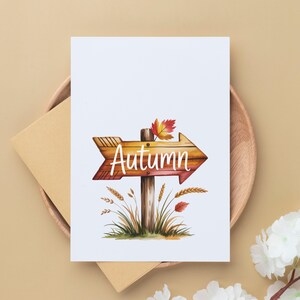 Pode incluir: Um cart&atilde;o branco com uma ilustra&ccedil;&atilde;o de uma placa de seta de madeira que diz "Autumn" em escrita branca. A placa &eacute; marrom com uma folha vermelha e amarela em cima. O cart&atilde;o est&aacute; em um prato de madeira com um envelope marrom.
