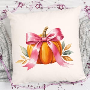 Pode incluir: Uma almofada quadrada decorativa com uma ilustra&ccedil;&atilde;o em aquarela de uma ab&oacute;bora laranja com um la&ccedil;o rosa. A ab&oacute;bora &eacute; adornada com folhas verdes e marrons. A almofada est&aacute; em um fundo branco.