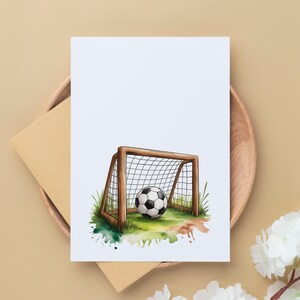 May include: A watercolor illustration of a soccer ball in a goal on a greeting card. The goal is brown with a white net, set on green grass with colorful paint splatters. The card is on a wooden plate with an envelope.