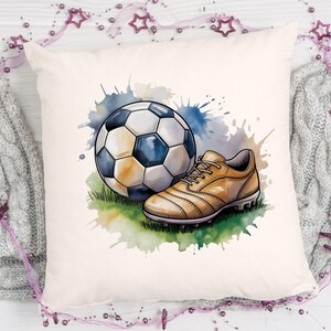 May include: A decorative pillow featuring a watercolor illustration of a soccer ball and a soccer cleat. The ball is white and black, and the cleat is tan. The background has blue and green watercolor splashes.