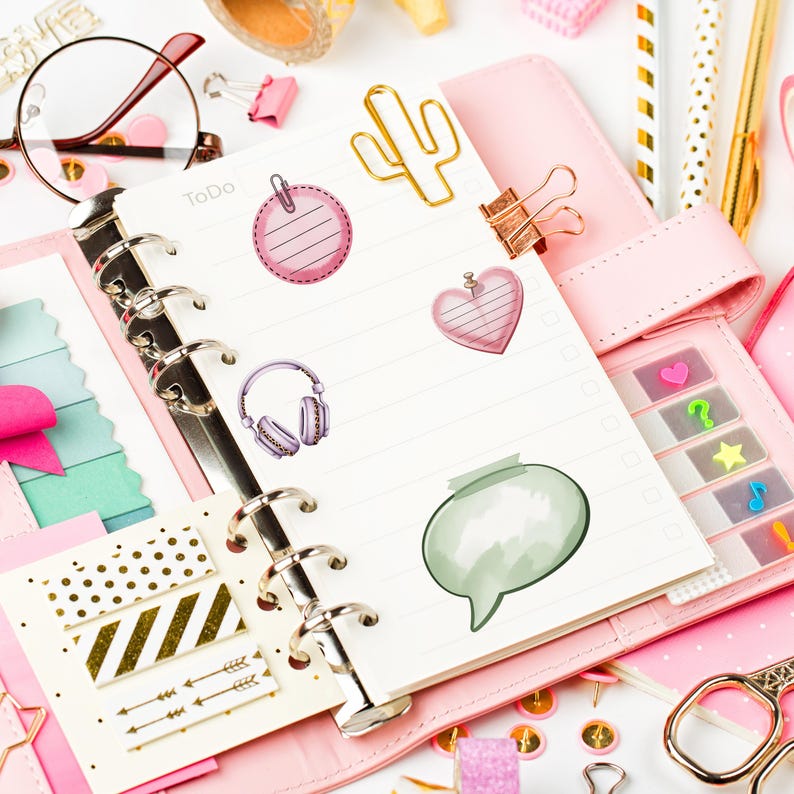 May include: A pink planner with a To Do list, decorated with stickers of a round tag, heart, headphones, and speech bubble. Gold cactus and paperclip accents, along with pens and scissors, complete the stationery theme.