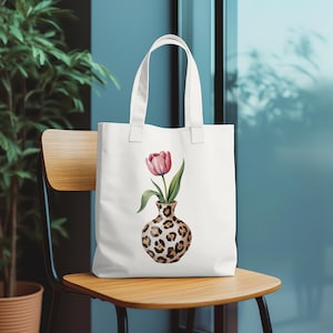 May include: White tote bag featuring a watercolor illustration of a pink tulip in a leopard print vase. The bag has a simple design with two handles and is sitting on a wooden chair.