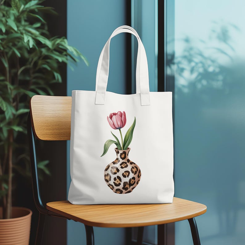 May include: White tote bag featuring a watercolor illustration of a pink tulip in a leopard print vase. The bag has a simple design with two handles and is sitting on a wooden chair.