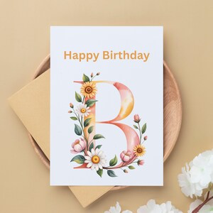 May include: A white greeting card with the words "Happy Birthday" in orange. The card features a large, floral-decorated letter "B" in shades of orange and pink, surrounded by sunflowers, daisies, and greenery. The card is on a wooden plate with a brown envelope.