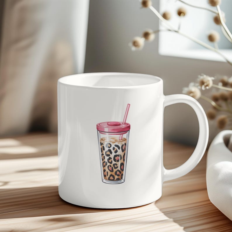 May include: White ceramic mug featuring a watercolor illustration of a leopard print iced coffee drink with a pink lid and straw. The mug has a large handle and is set on a wooden surface.