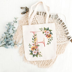 May include: A cream-colored tote bag featuring a floral design around the letter "F". The floral arrangement includes sunflowers, daisies, and green leaves. The bag has cream-colored handles and is displayed on a white surface.
