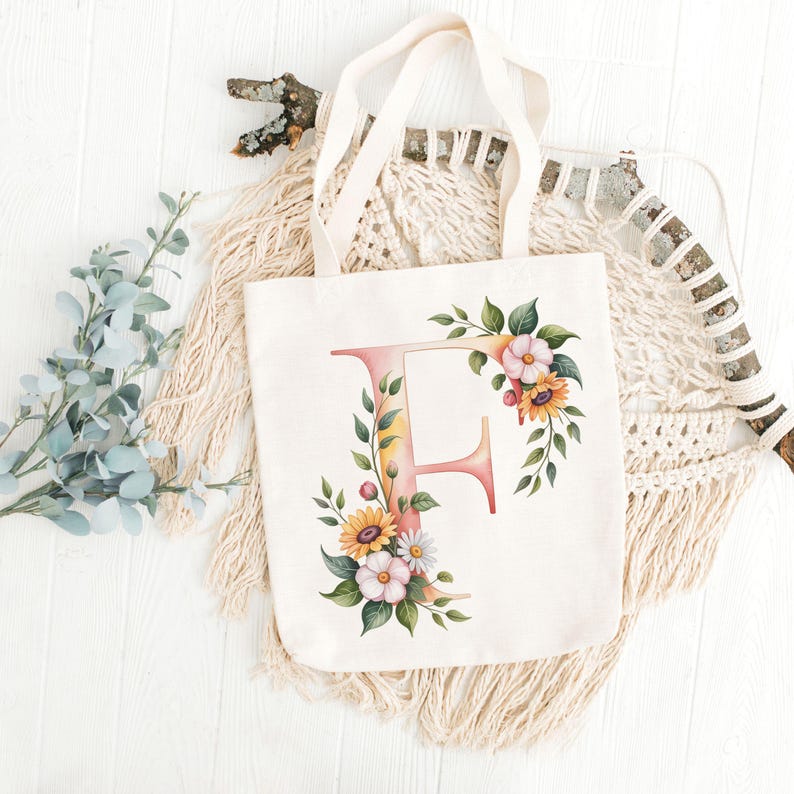 May include: A cream-colored tote bag featuring a floral design around the letter "F". The floral arrangement includes sunflowers, daisies, and green leaves. The bag has cream-colored handles and is displayed on a white surface.