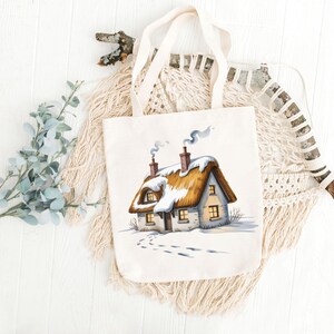 May include: A cream-colored tote bag featuring a watercolor illustration of a snow-covered cottage with a thatched roof. The cottage has two chimneys with smoke, and footprints lead to the door. The bag has two cream-colored handles.