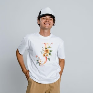 May include: White t-shirt featuring a watercolor floral letter "J" design. The design includes sunflowers, daisies, and other flowers in shades of yellow, orange, and pink. The shirt is paired with a black and white trucker hat.