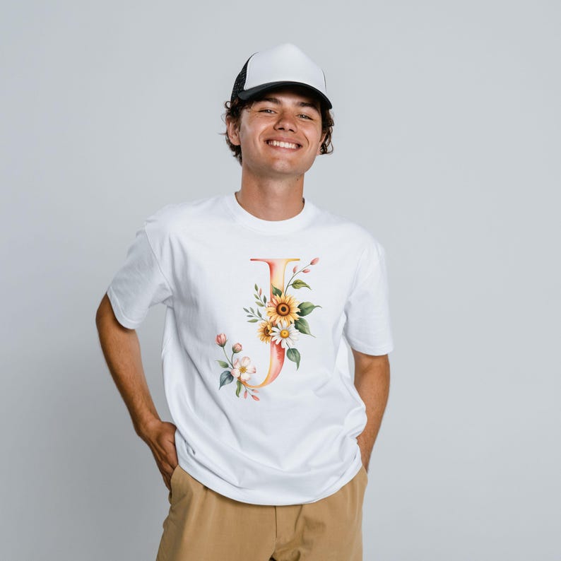 May include: White t-shirt featuring a watercolor floral letter "J" design. The design includes sunflowers, daisies, and other flowers in shades of yellow, orange, and pink. The shirt is paired with a black and white trucker hat.